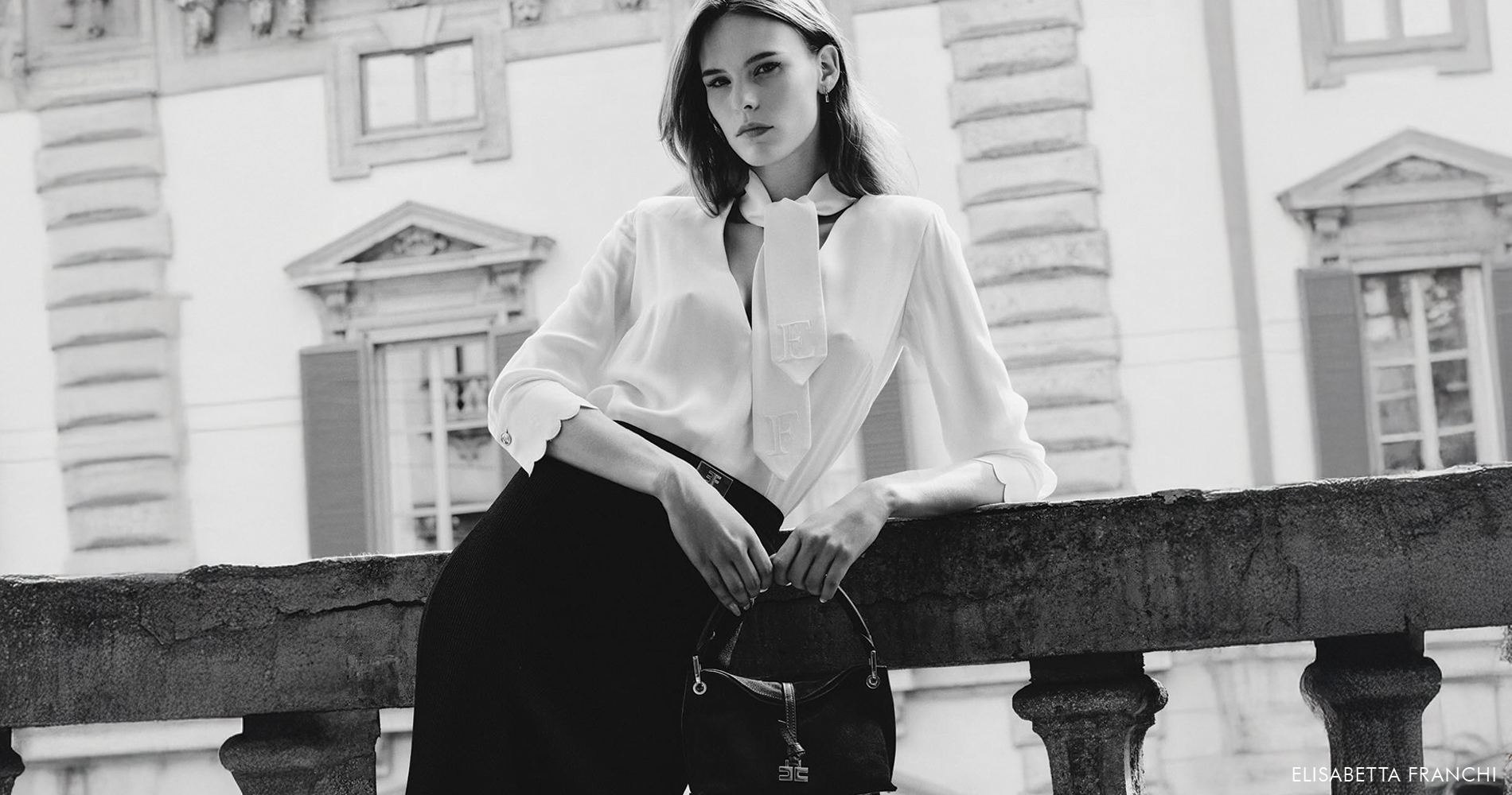 An elegant woman in a white blouse and black skirt confidently poses by a stone railing of a historic Italian building, embodying modern Italian elegance.