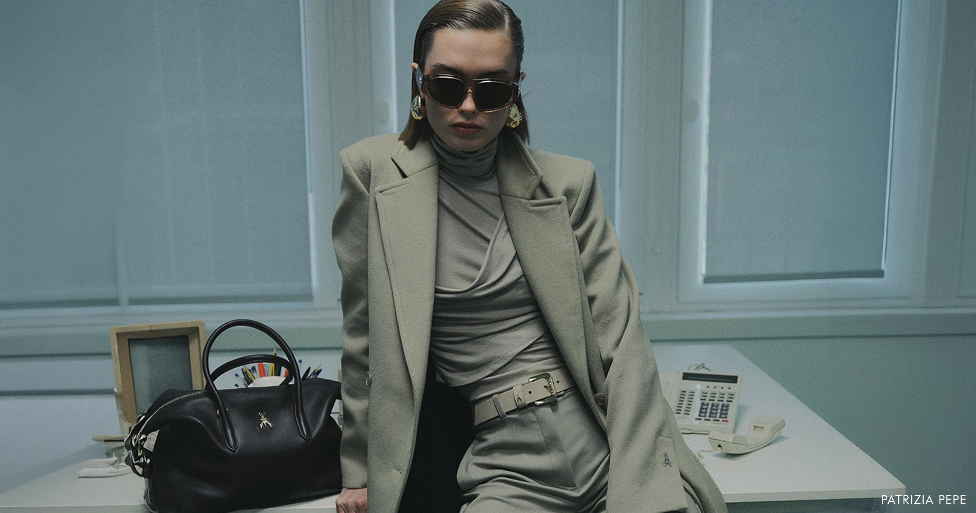 A woman in a modern monochrome outfit with an oversized blazer sits confidently in an office setting, reflecting urban Italian elegance.