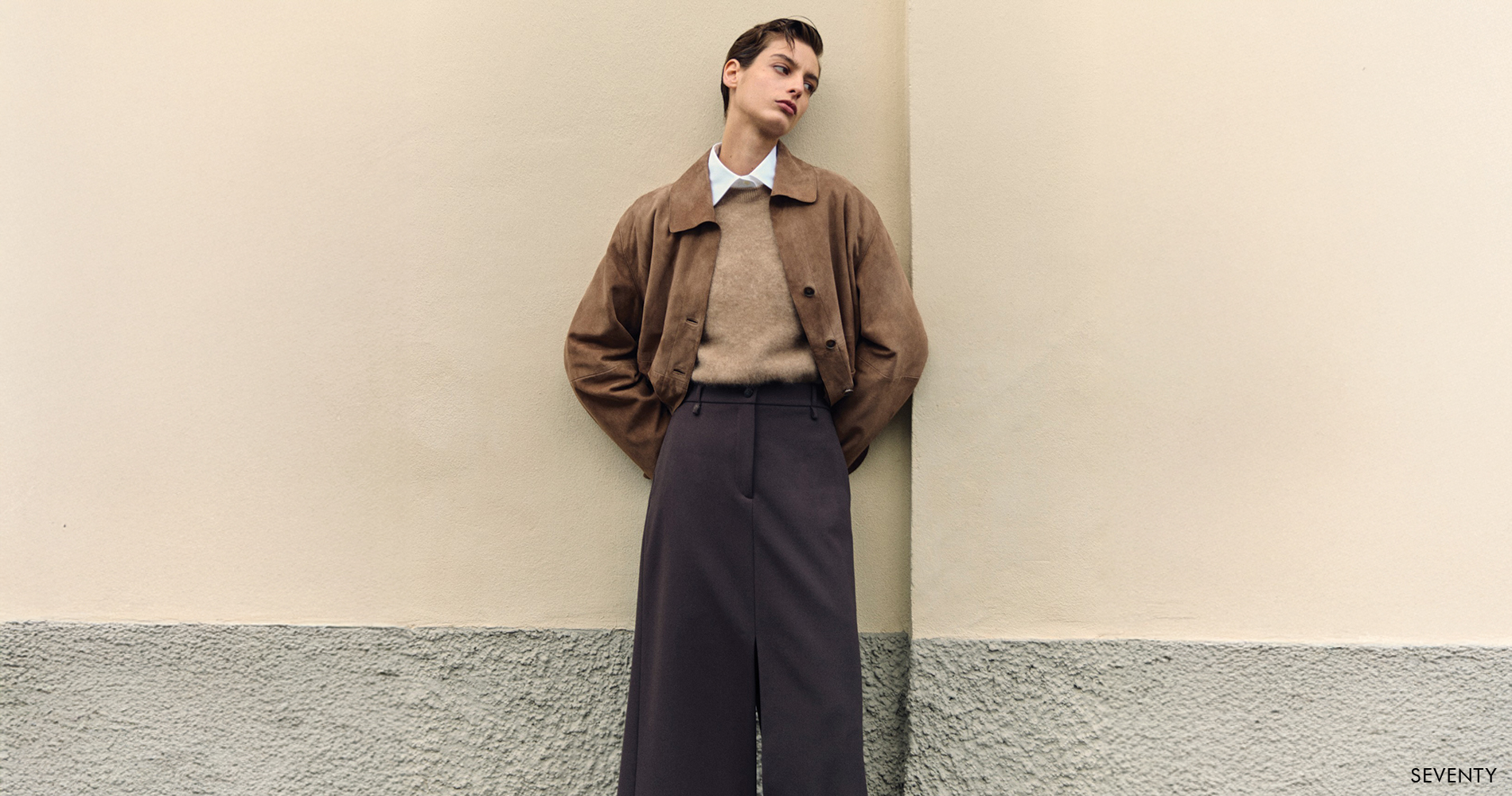 A woman in a soft brown jacket, wool sweater and tailored trousers stands against a light wall, embodying refined Italian elegance in warm earthy tones.