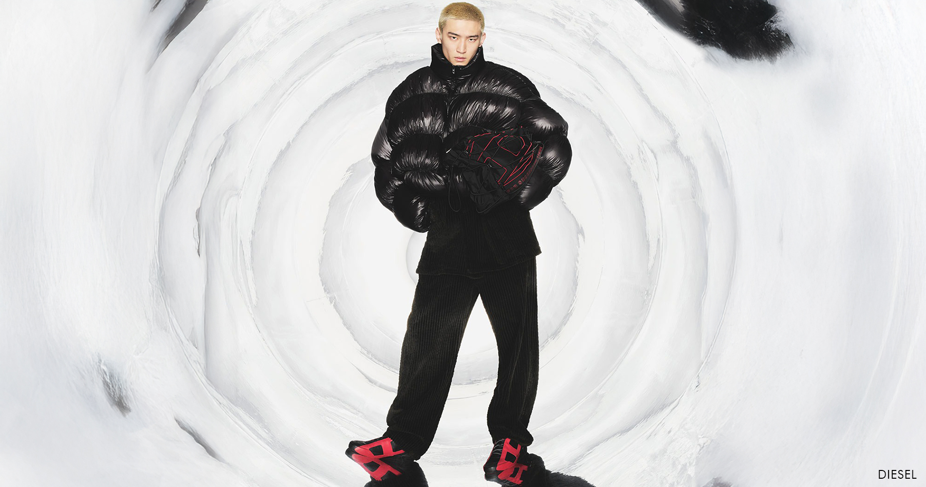 Moški v črni puhovki in žametnih hlačah pozira pred svetlim ozadjem v sodobnem italijanskem slogu.