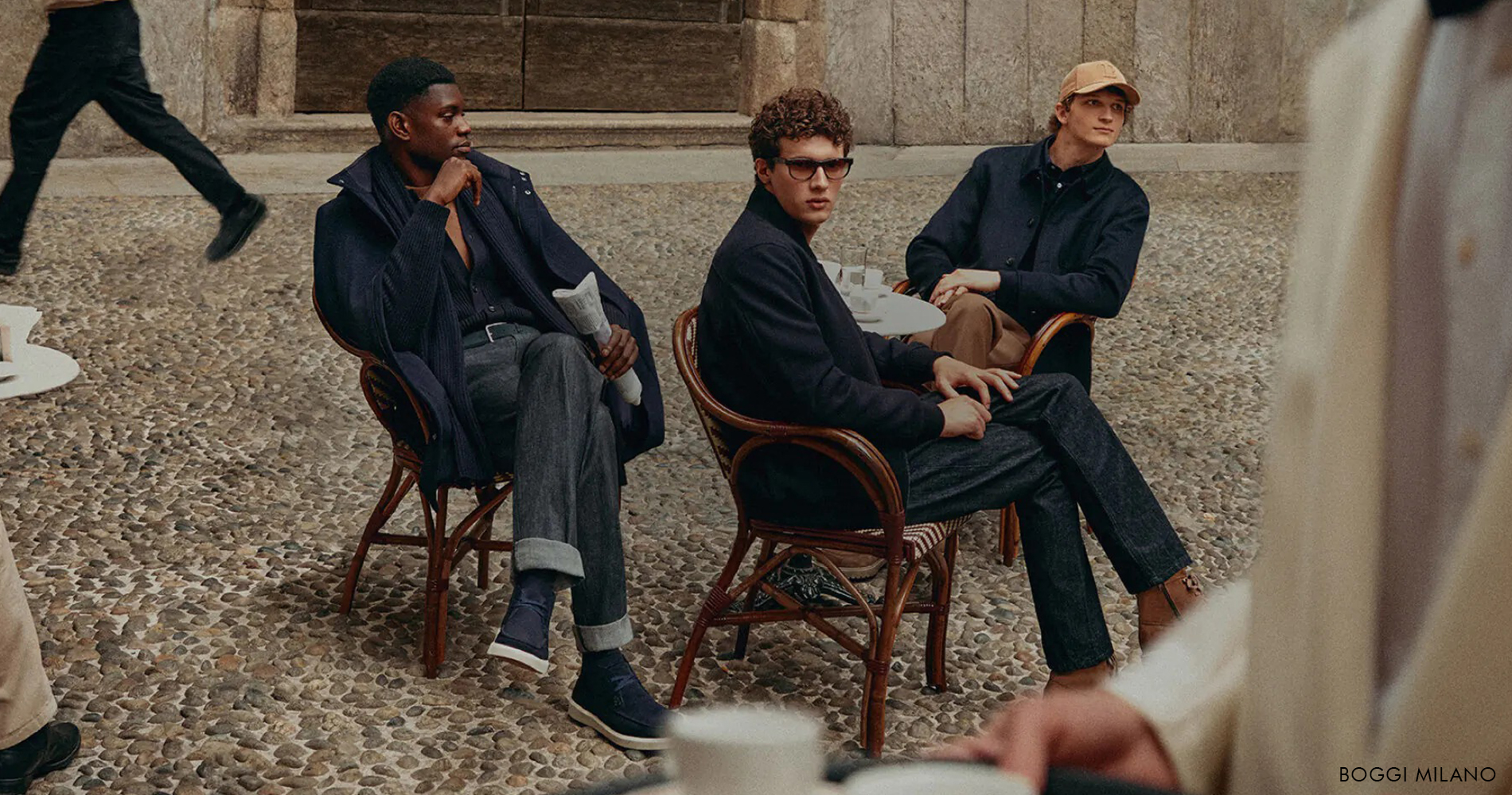 Three men in relaxed dark outfits sit at a café terrace, captured in a moment of contemporary Italian street style.