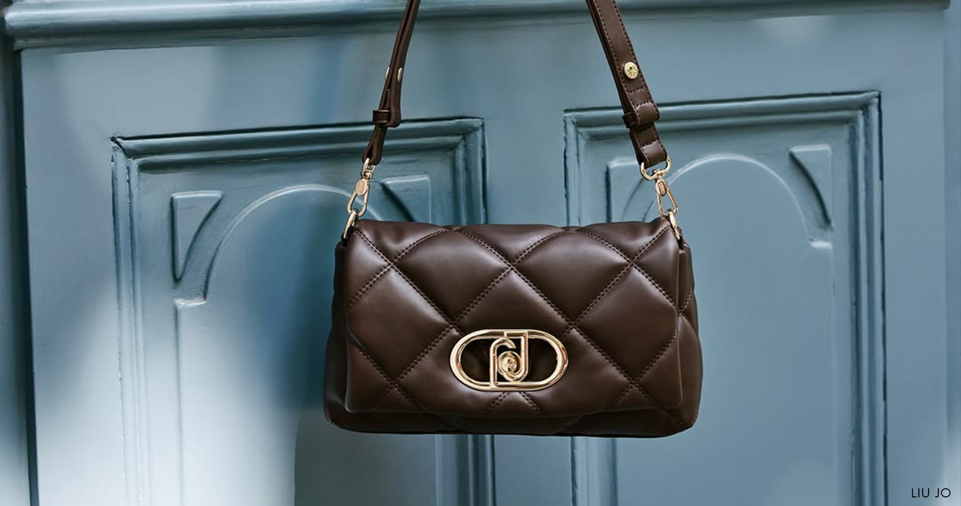 An elegant brown quilted handbag with a gold clasp hangs from an ornate metal handle against a blue wooden surface.