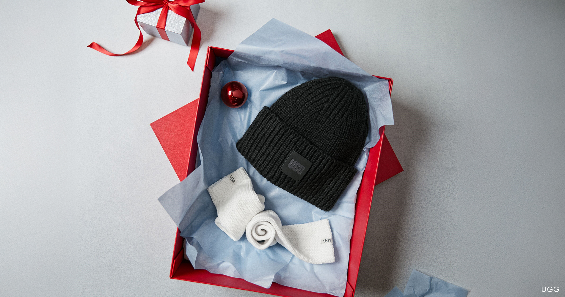 Praznični darilni set UGG, ki vključuje črno pleteno kapo in bele tople nogavice, elegantno predstavljen v rdeči darilni škatli.