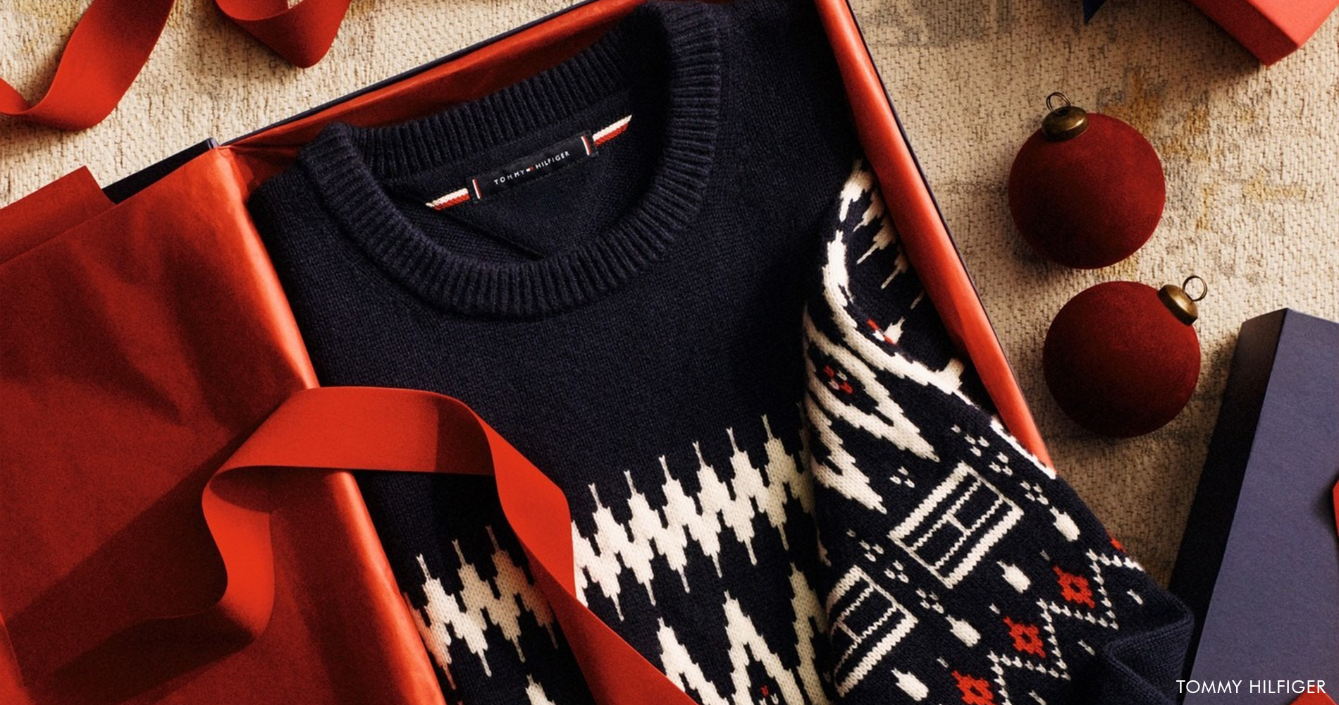 Elegantno zapakiran pleten pulover s prazničnim vzorcem v darilni škatli, obdan z rdečimi trakovi, okrasnimi kroglicami in prazničnimi darili, ustvarja topel božični ambient.