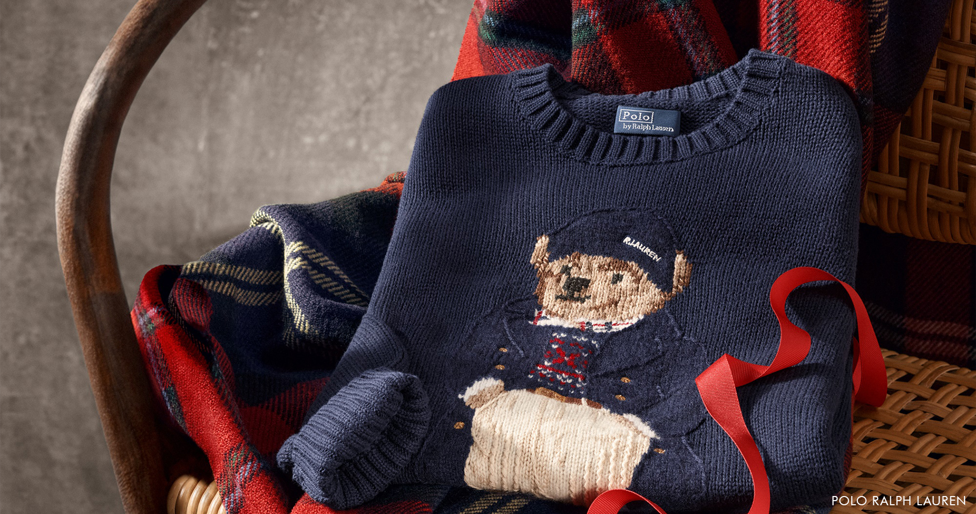 Topel pleten pulover Polo Ralph Lauren z ikoničnim medvedjim motivom, položen na mehko karirasto odejo v prazničnem zimskem vzdušju.