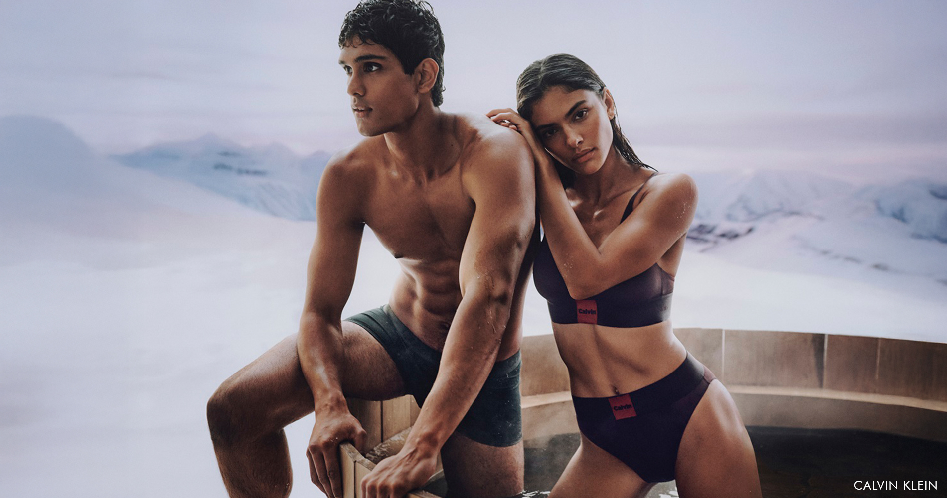 Eleganten par v jacuzziju na prostem, oblečen v črne in temno vijolične kose spodnjega perila Calvin Klein, s snežno pokrajino v ozadju.