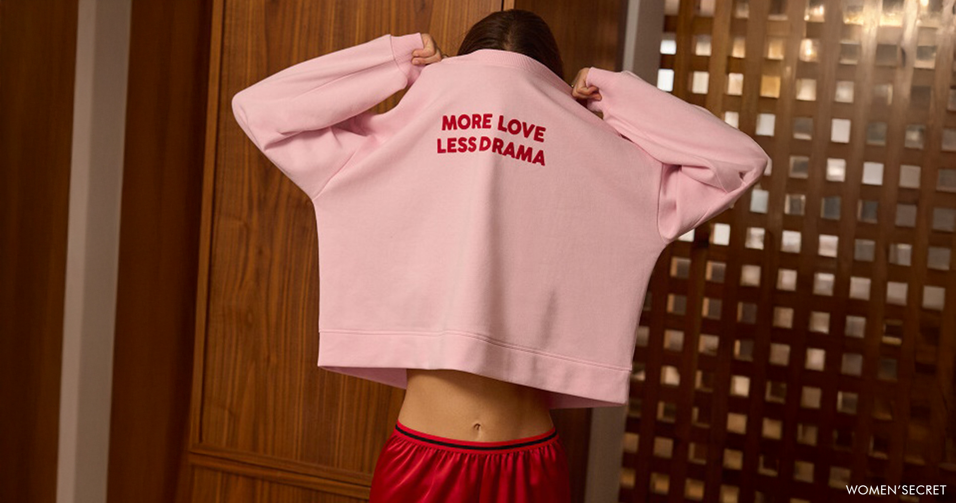 Ženska v rožnatem puloverju in rdečih kratkih hlačah, sproščen domač videz z napisom More love less drama.