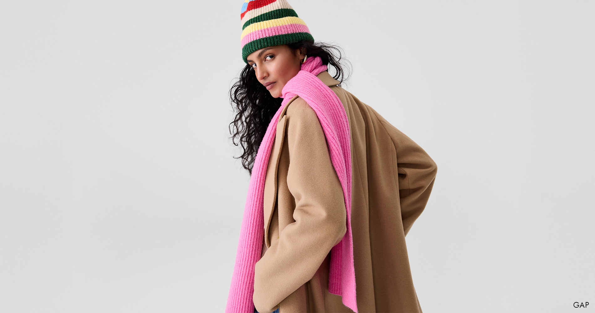 Ženska v bež plašču nosi pisano pleteno kapo in rožnat šal, ki z živahnimi barvami popestrita zimski videz.