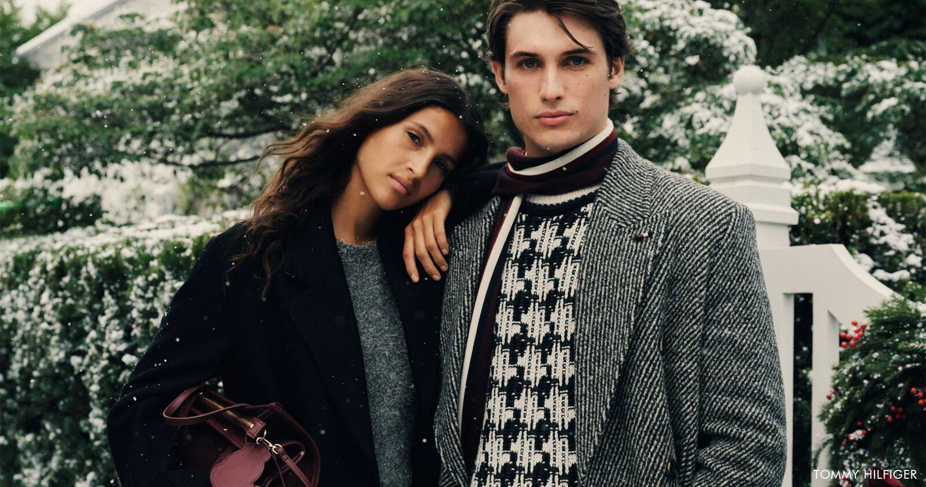 Elegantna ženska in moški v toplih zimskih plaščih in pleteninah pozirata na prostem med rahlim sneženjem ter utelešata brezčasno zimsko eleganco in udobje.