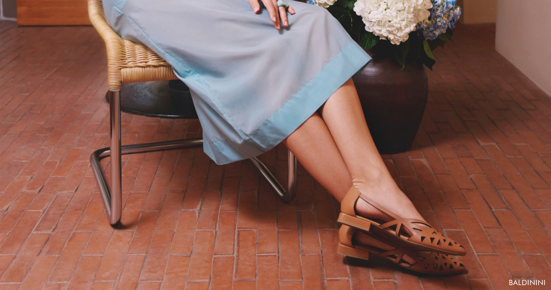 Ženska v svetlo modrem monochrome outfitu sedi na stolu iz rafije, obdana s cvetlicami, obuta v rjave balerinke.