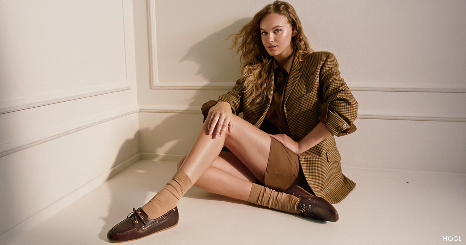 Ženska v rjavem suknjiču, kratkih hlačah in rjavih čevljih sproščeno sedi na bež tleh.