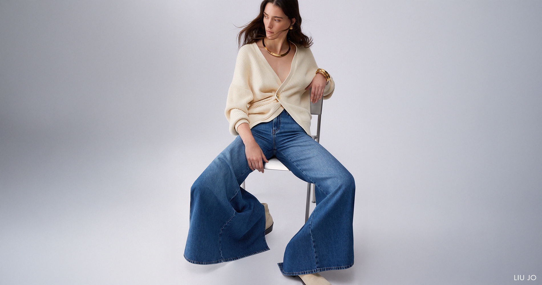 Ženska v jeans hlačah na zvonec in bež pletenini sproščeno sedi na belem stolu.