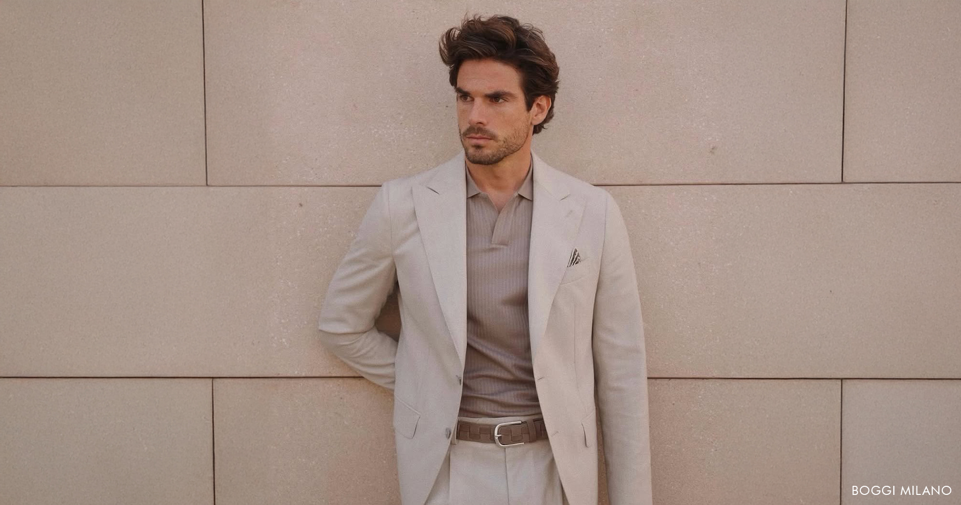 A man in a beige suit casually leaning against a beige wall, exuding elegance and confidence.