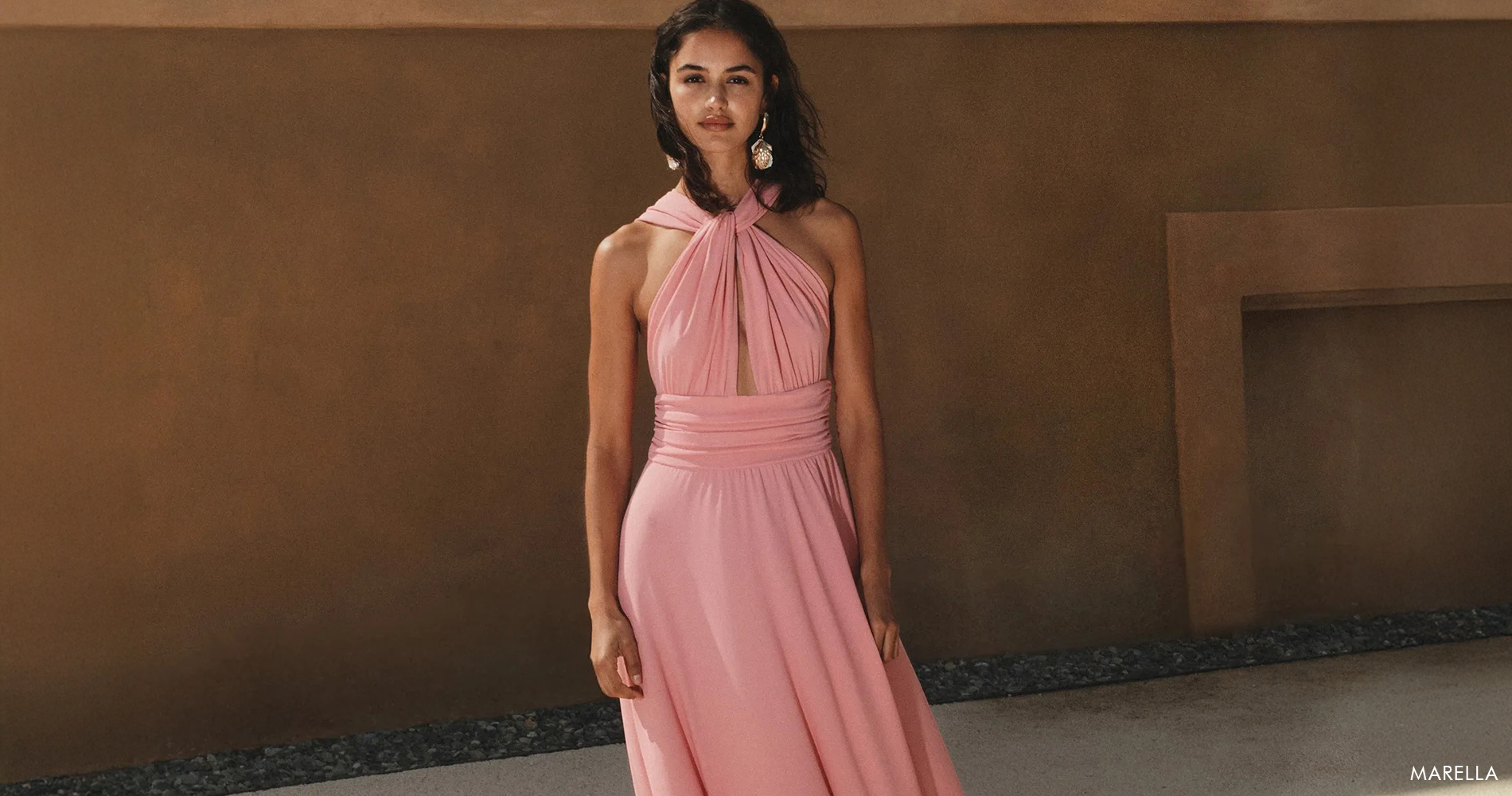 A smiling young woman in a soft pink elegant flowing dress against a brown background.