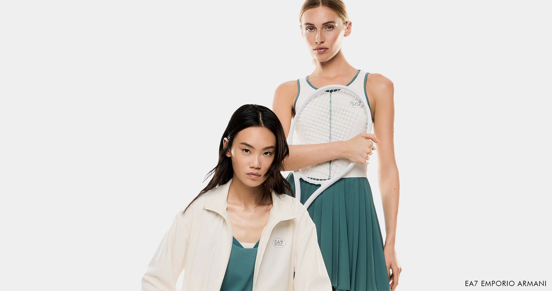 Two women in sporty-elegant outfits stand against a light background; one holds a tennis racket and wears a pleated skirt, while the other wears a lightweight jacket and a matching top.