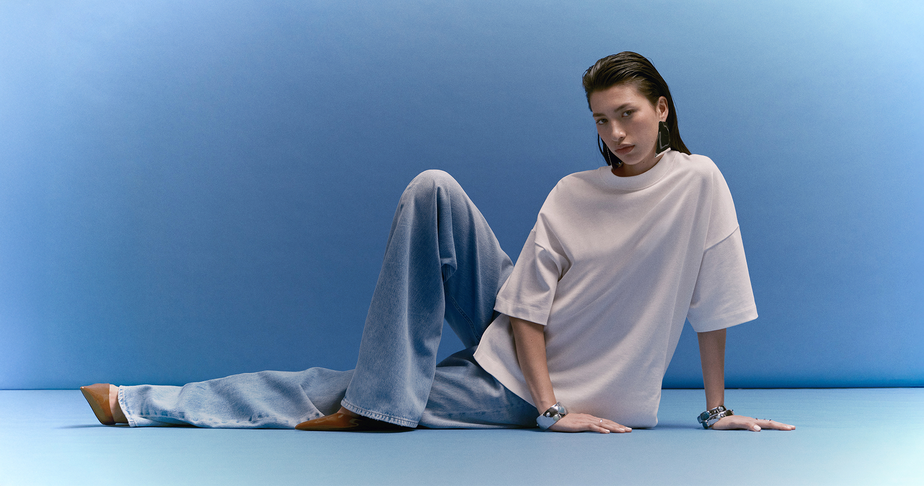 Model v beli majici in širokih svetlo modrih kavbojkah pozira na modrem ozadju. Minimalističen styling in sproščena silhueta poudarjata sodoben videz.