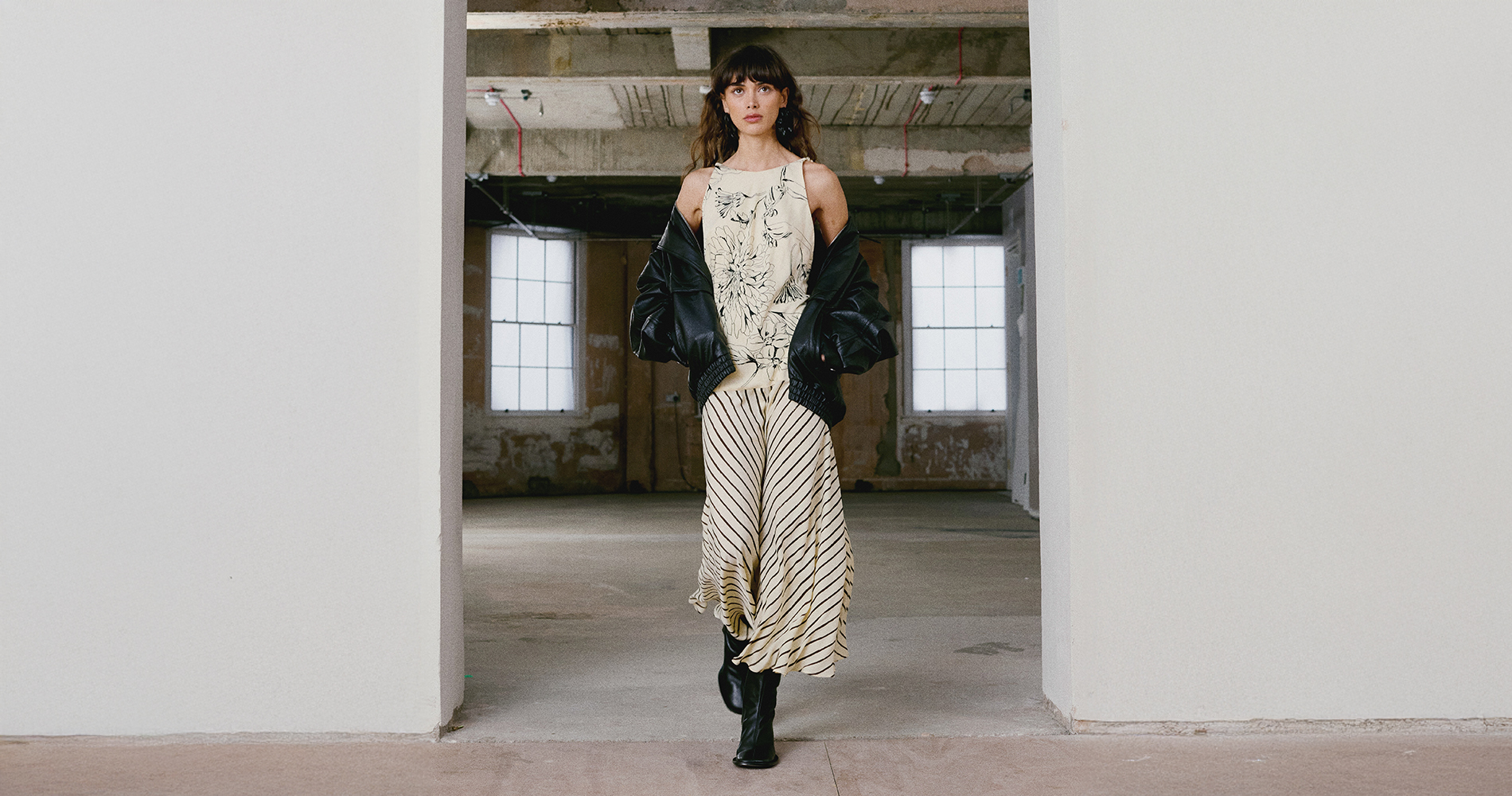 Model v lahkotni vzorčasti obleki in črni usnjeni jakni hodi skozi minimalističen industrijski prostor. Kontrast med tekočo silhueto in robustnimi detajli ustvarja sodoben, urban videz.