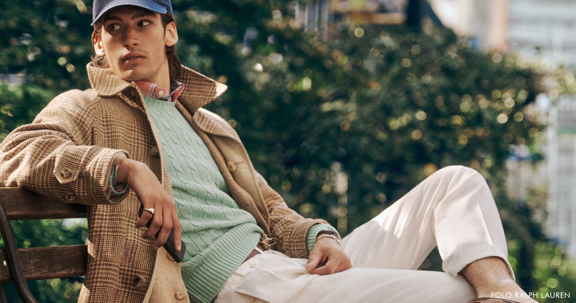 A fashionably dressed man sits casually on a bench in a park, with a modern, urban atmosphere.