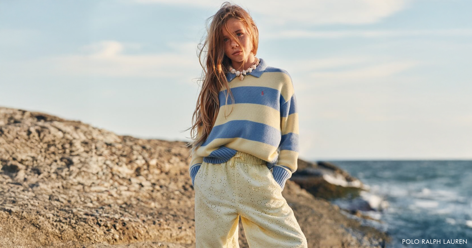 A girl stands by the sea, wearing a striped sweater in pastel colors and pastel yellow trousers.