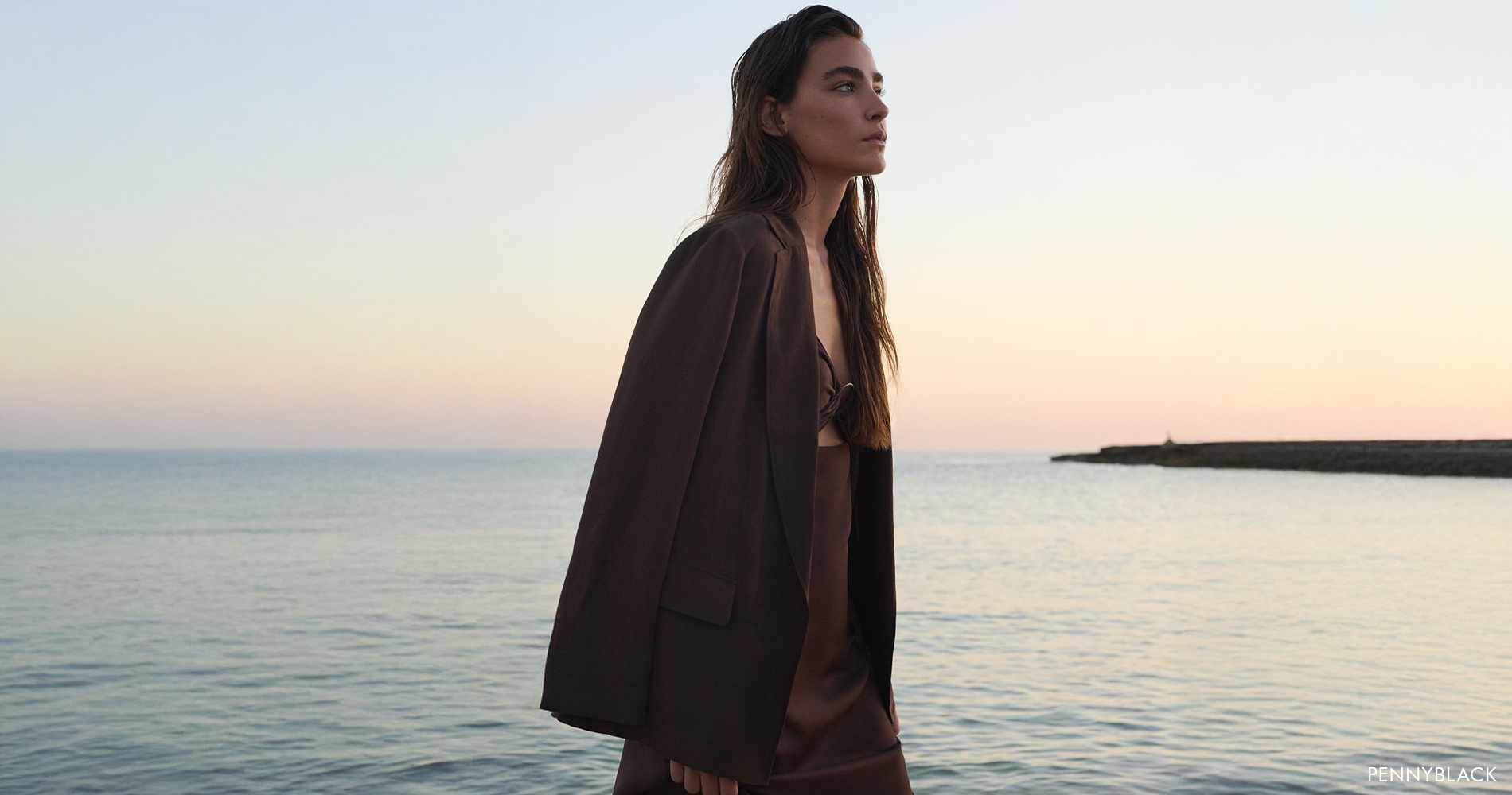 A woman in a monochrome chocolate brown look stands thoughtfully by the sea, with a sunset in the background.